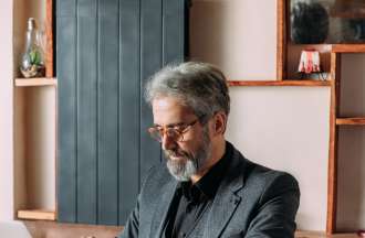 [Featured image] A person with gray hair and a beard wearing a gray sports jacket, black shirt, and glasses is sitting at a desk working on a laptop. 