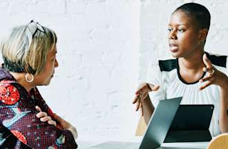 [Featured image] A female business analyst interviews a female employee. Both have their laptops in front of them.