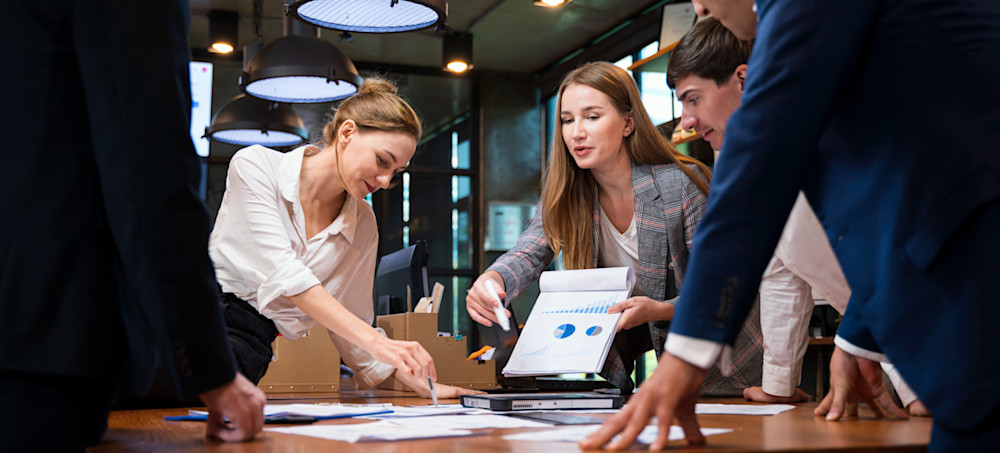 [Featured Text]: A group of people stand around a table to discuss the types of marketing they plan to use in an upcoming campaign.
