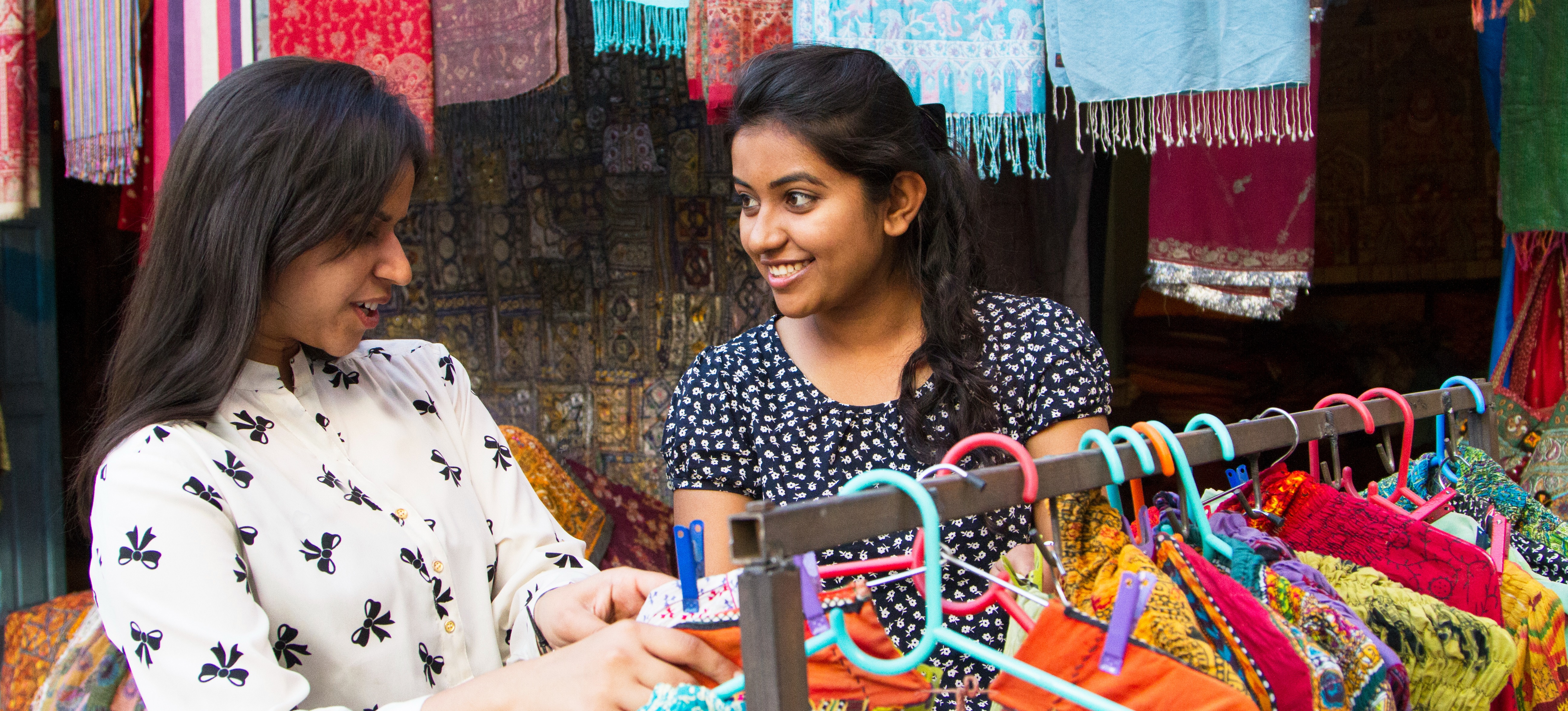 [Featured Image] A smiling entrepreneur who makes clothing talks with a customer inside her shop as they both look through colorful clothing.