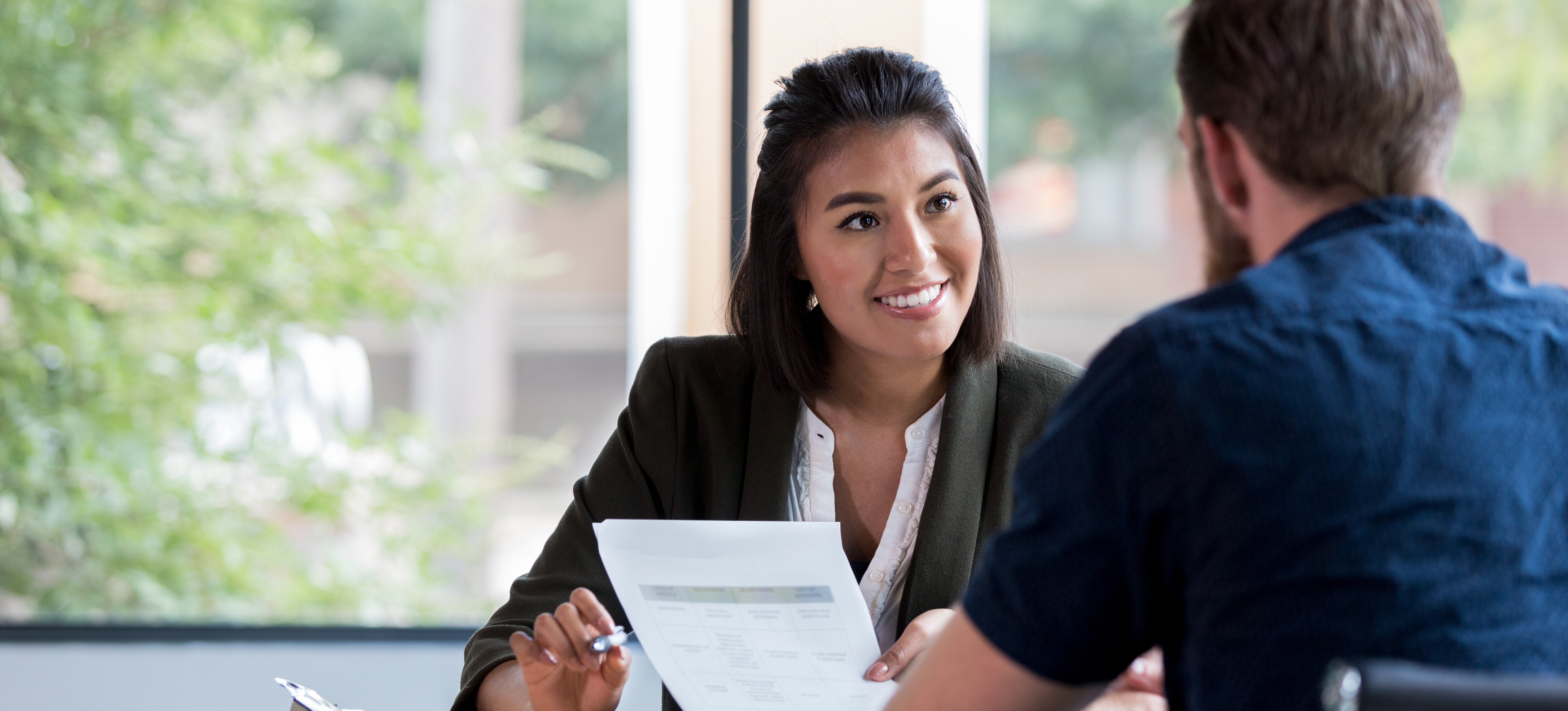 [Featured Image]: A smiling HR manager holds a resume and talks with a potential employee while they are both sitting at a table in a sunny office.
