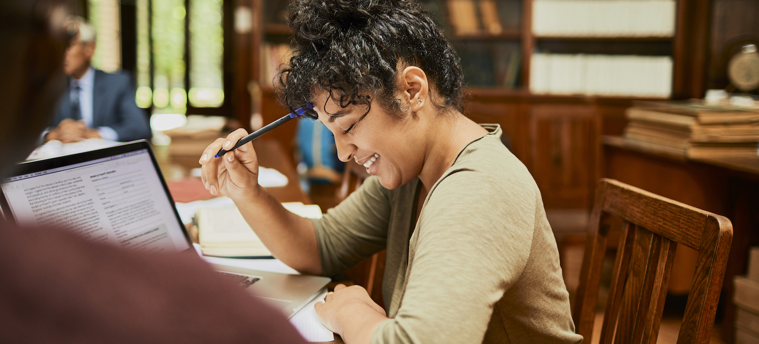 [Featured image] A student sits in their school library with a laptop open, smiling while they study for a college class.