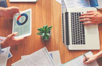 [Featured image] Several coworkers share data analytics around a wooden table covered in laptops, tablets, and paperwork. 