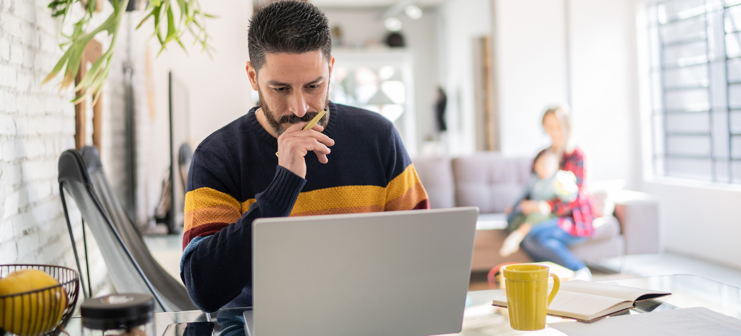 [Featured image] A person stares concernedly at their laptop while another person with a baby sits in the background. 