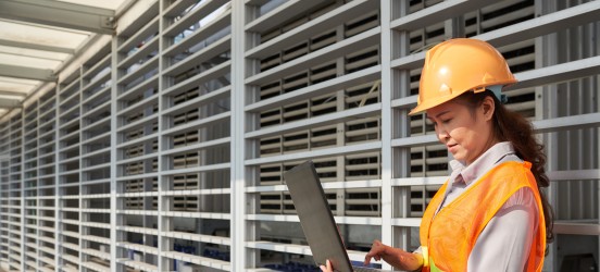 [Featured image] An electrical engineer in an orange helmet and safety vest reviews charts and information on the job site. 