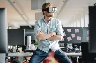[Featured Image] A VR worker tests virtual realty goggles at his desk.