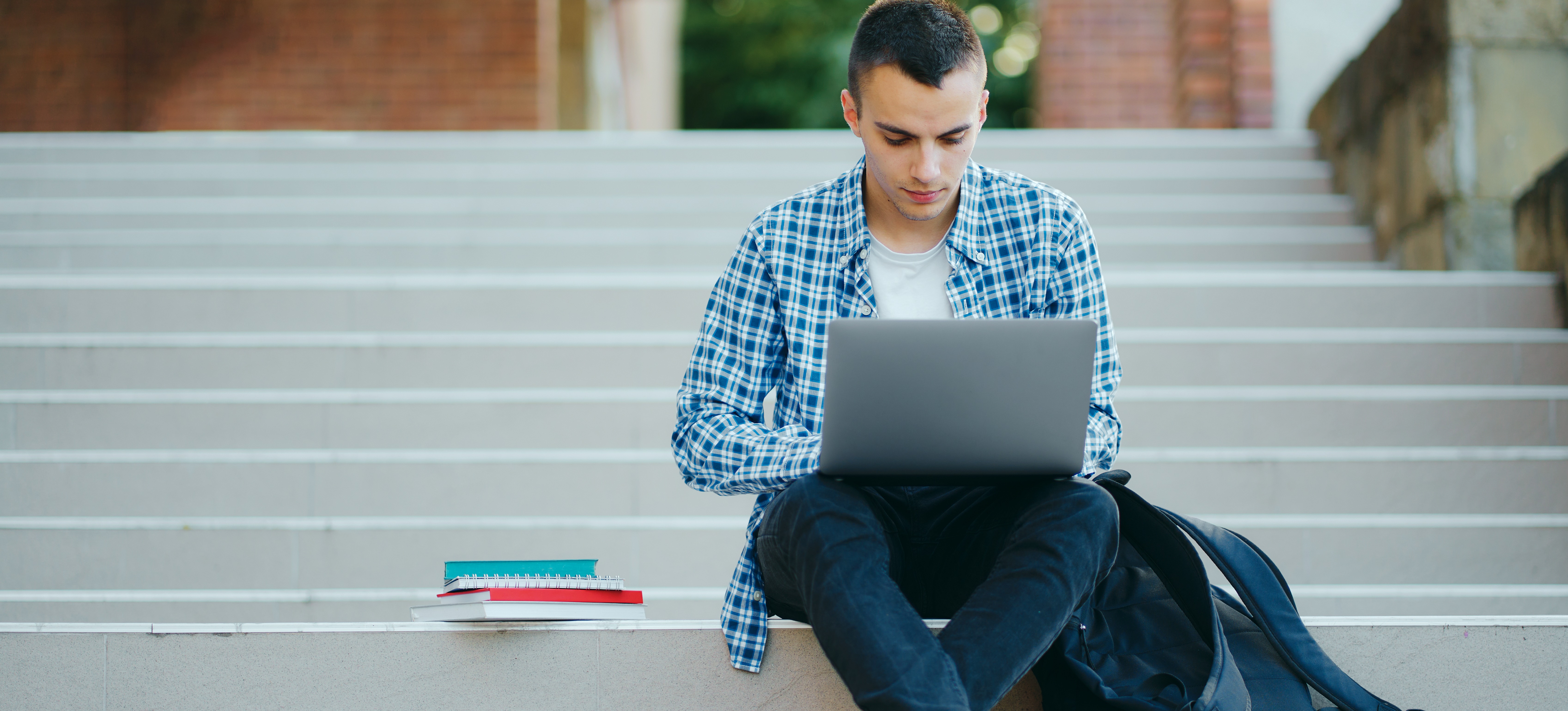 [Featured Image] A student sits on the steps on his college campus and uses a laptop to research whether he can get another bachelor's degree after graduating. 