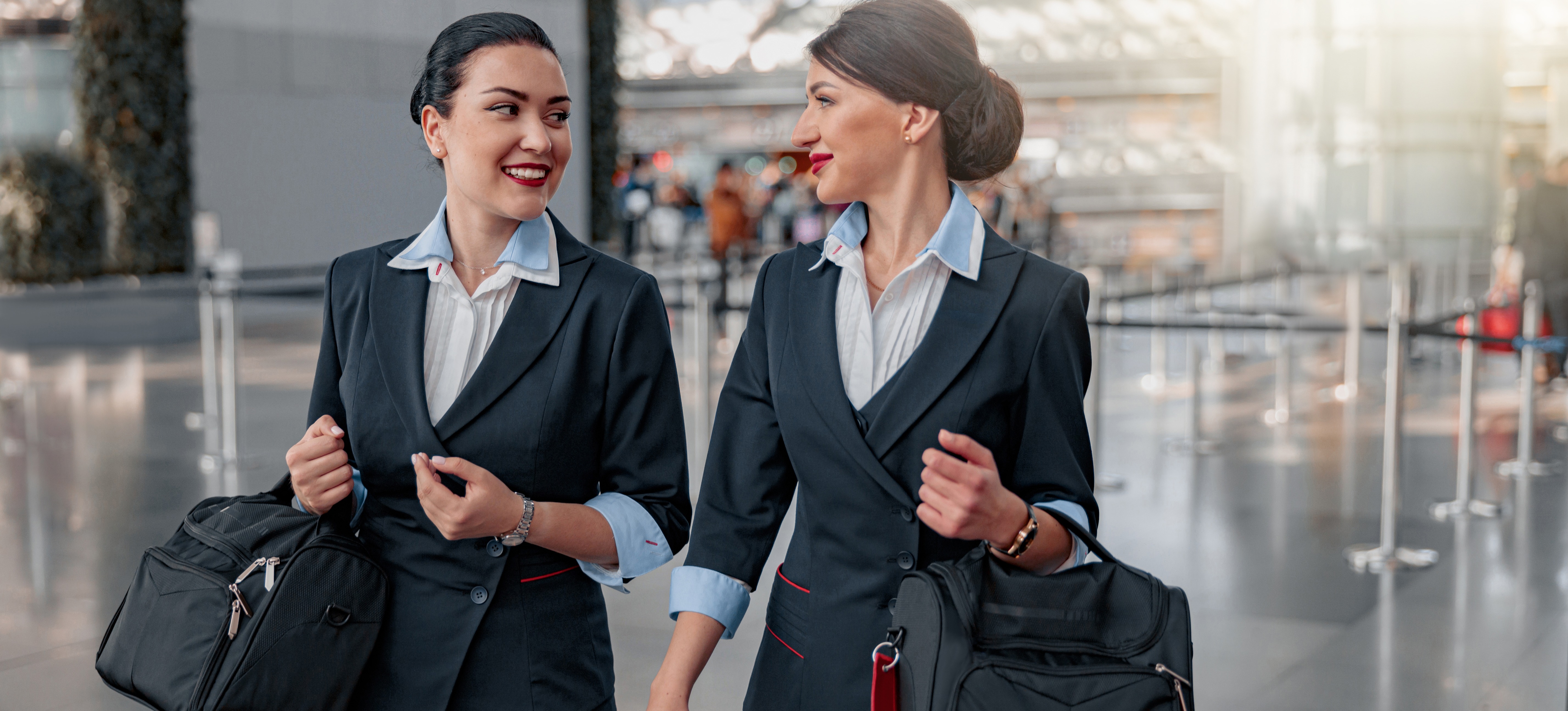 [Featured Image] Two flight attendants carry their bags and talk while walking through an airport.
