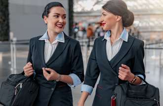 [Featured Image] Two flight attendants carry their bags and talk while walking through an airport.
