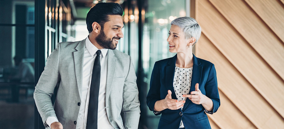 [Featured Image] Two colleagues dressed in professional attire smile and talk while walking through an office corridor as they discuss how to choose a career.
