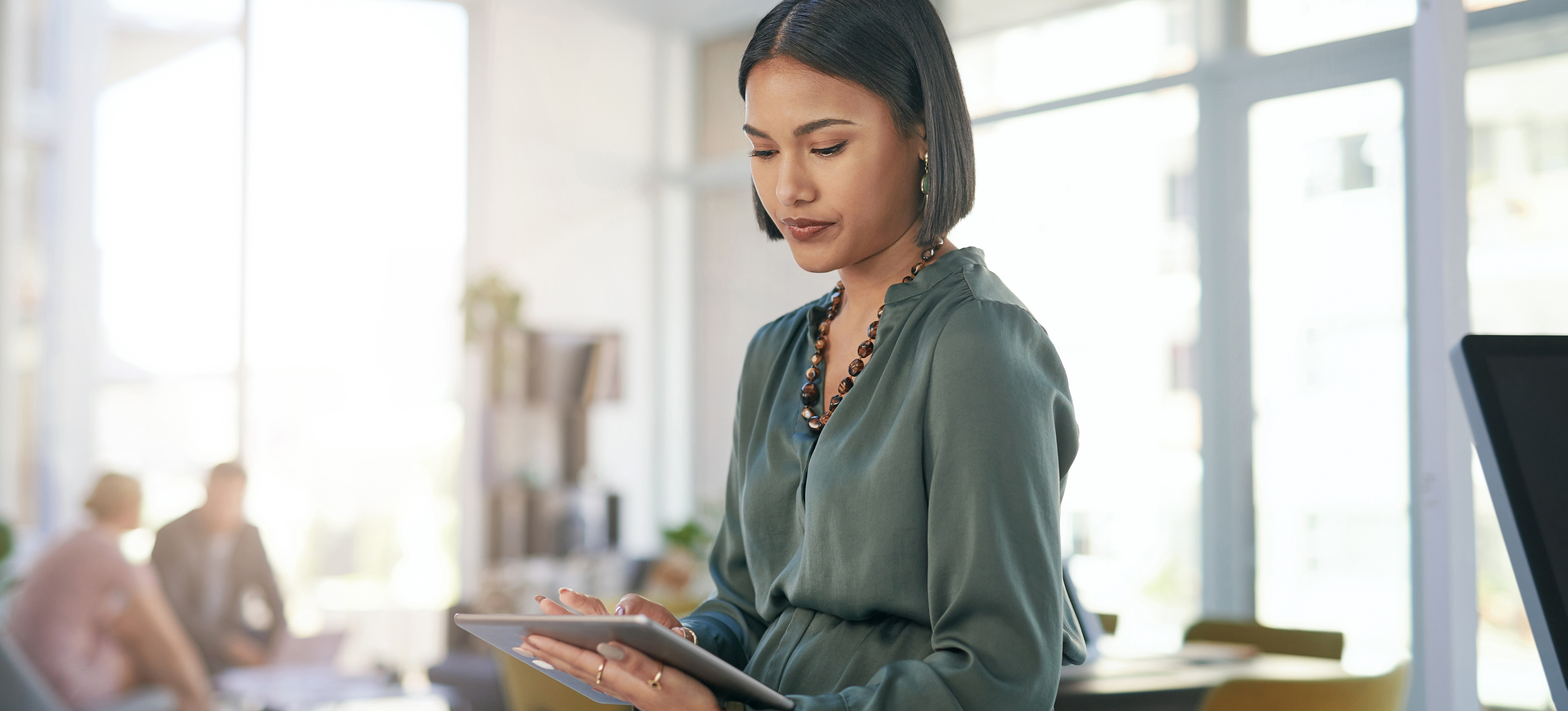 [Featured Image] In a bright office setting, a woman working for an enterprise architect salary looks at a tablet while she leans against the desk behind her.
