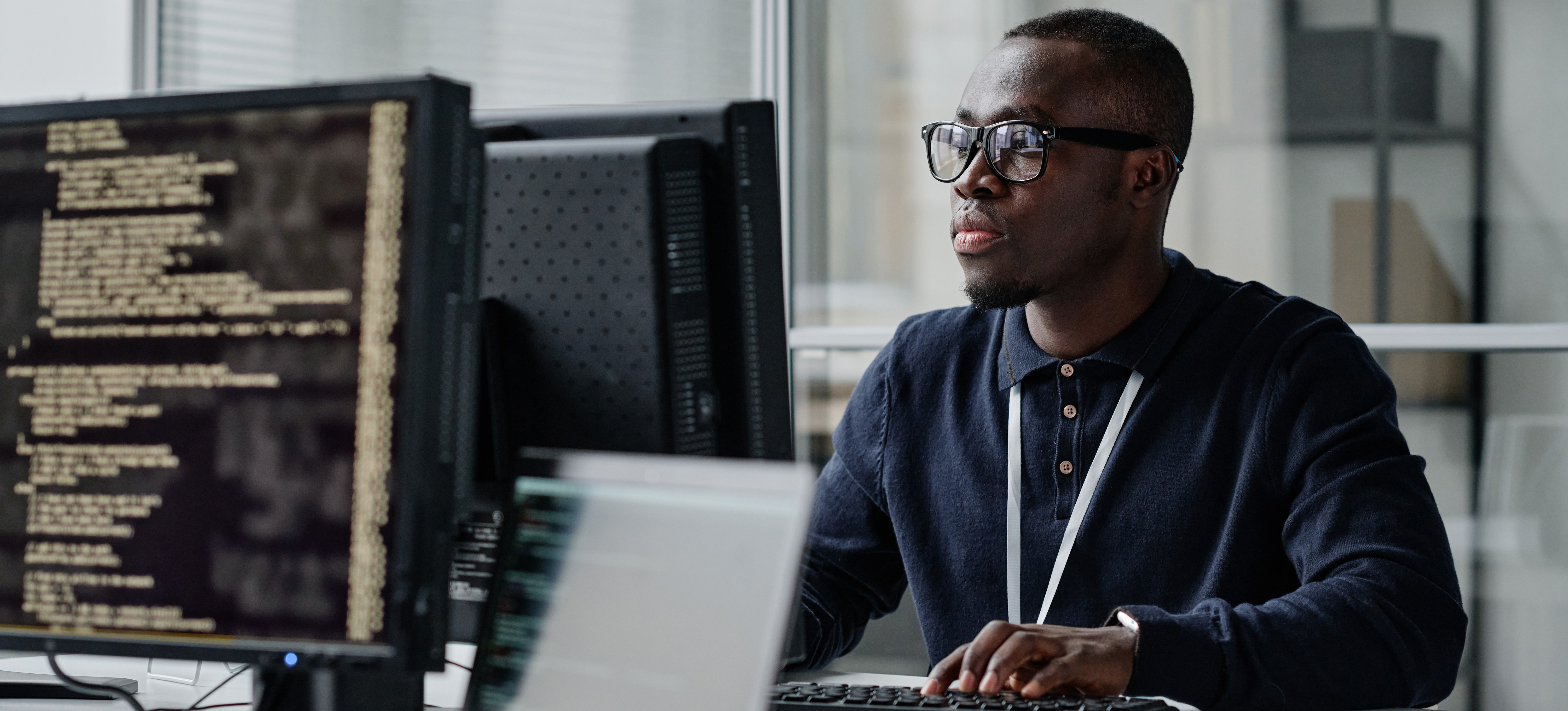 [Featured Image] A back-end developer in a blue shirt works at a computer in an open office space.