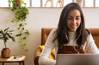 [Featured image] A concentrated, confident learner in a white shirt sits at the table and browses a laptop, and listens to music in a workspace.