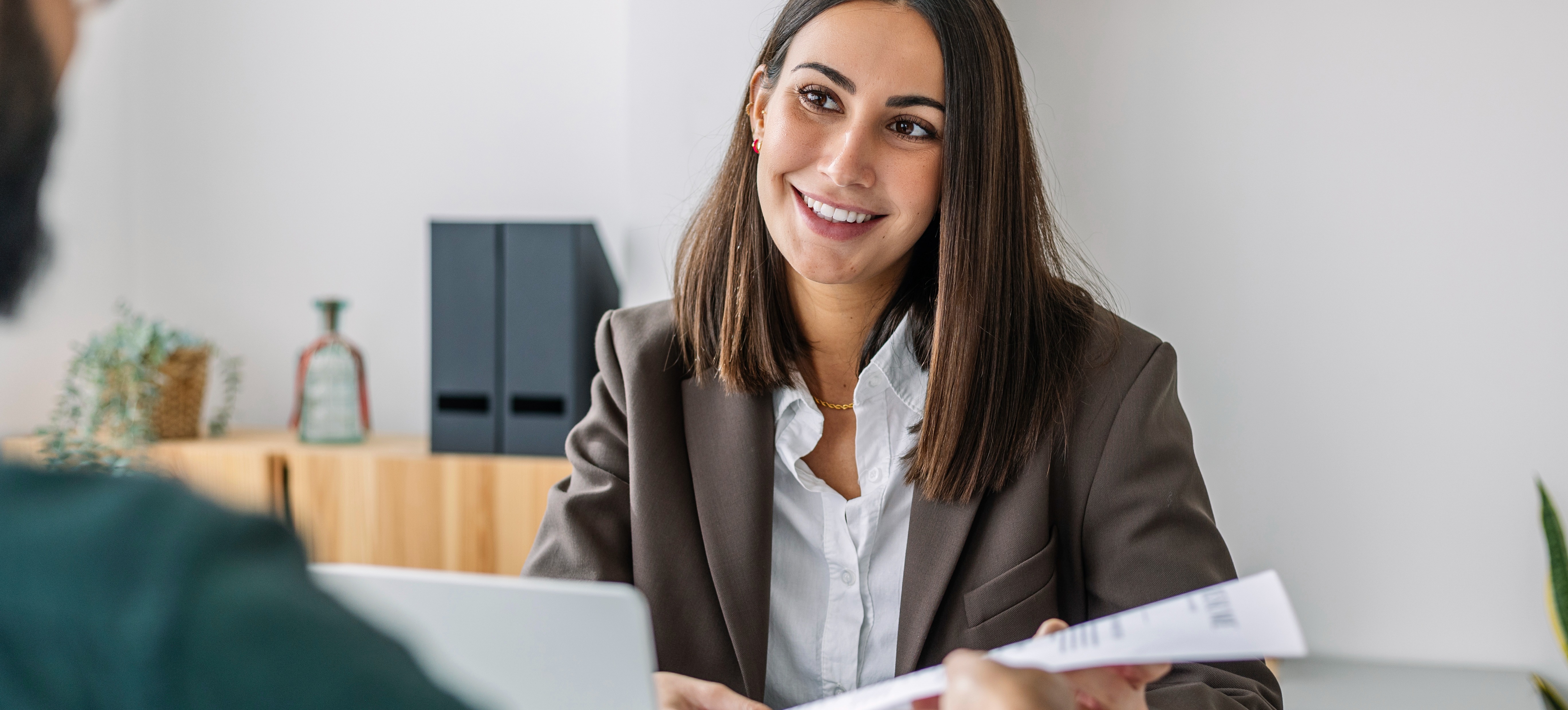 [Featured Image] A person hands their resume to a potential employer during a job interview and wonders if they are overqualified for a job. 