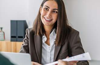 [Featured Image] A woman hands her resume to a potential employer during a job interview and wonders if she is overqualified for a job. 