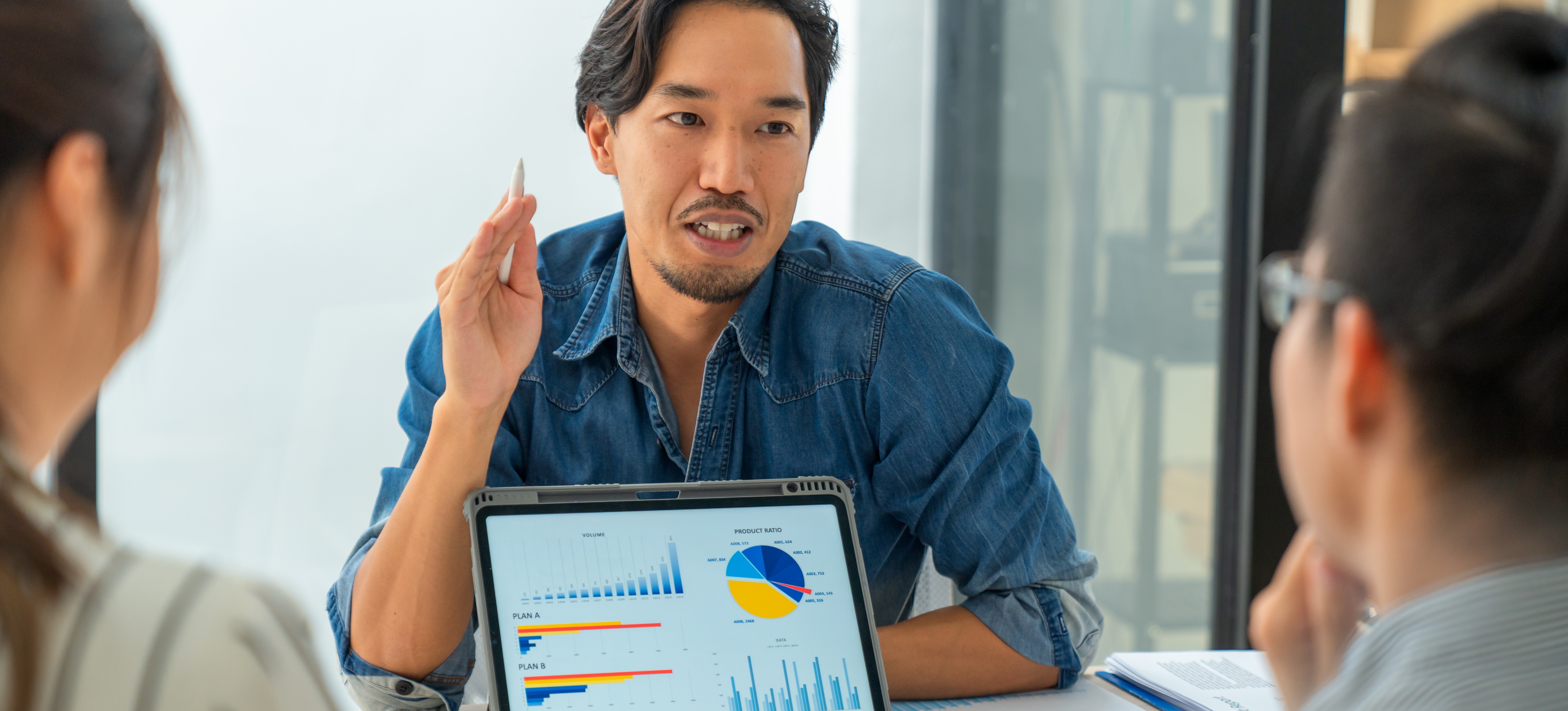 [Featured Image] A data analyst discusses visualizations generated by AI in analytics displayed on a tablet with two colleagues.