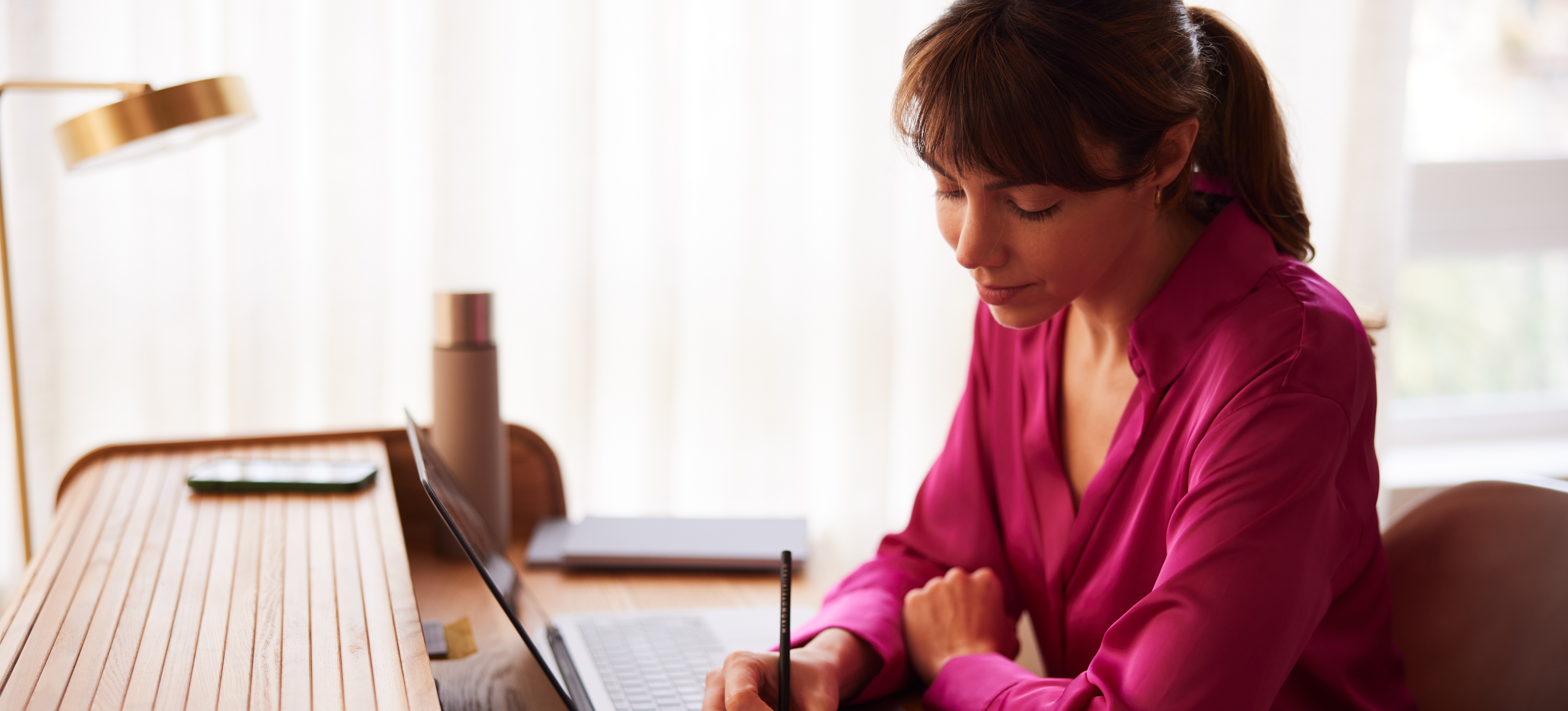 [Featured Image] A woman in a bright pink blouse sits at her desk, writing in a notebook and adding up how many work hours in a year.

