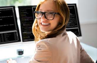 [Featured image] A woman sits in front of two computer monitors writing code in a programming language.