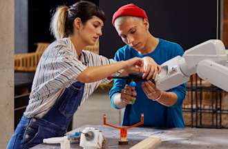 [Featured Image] Two robotics engineers work together on a robotic arm in a warehouse.