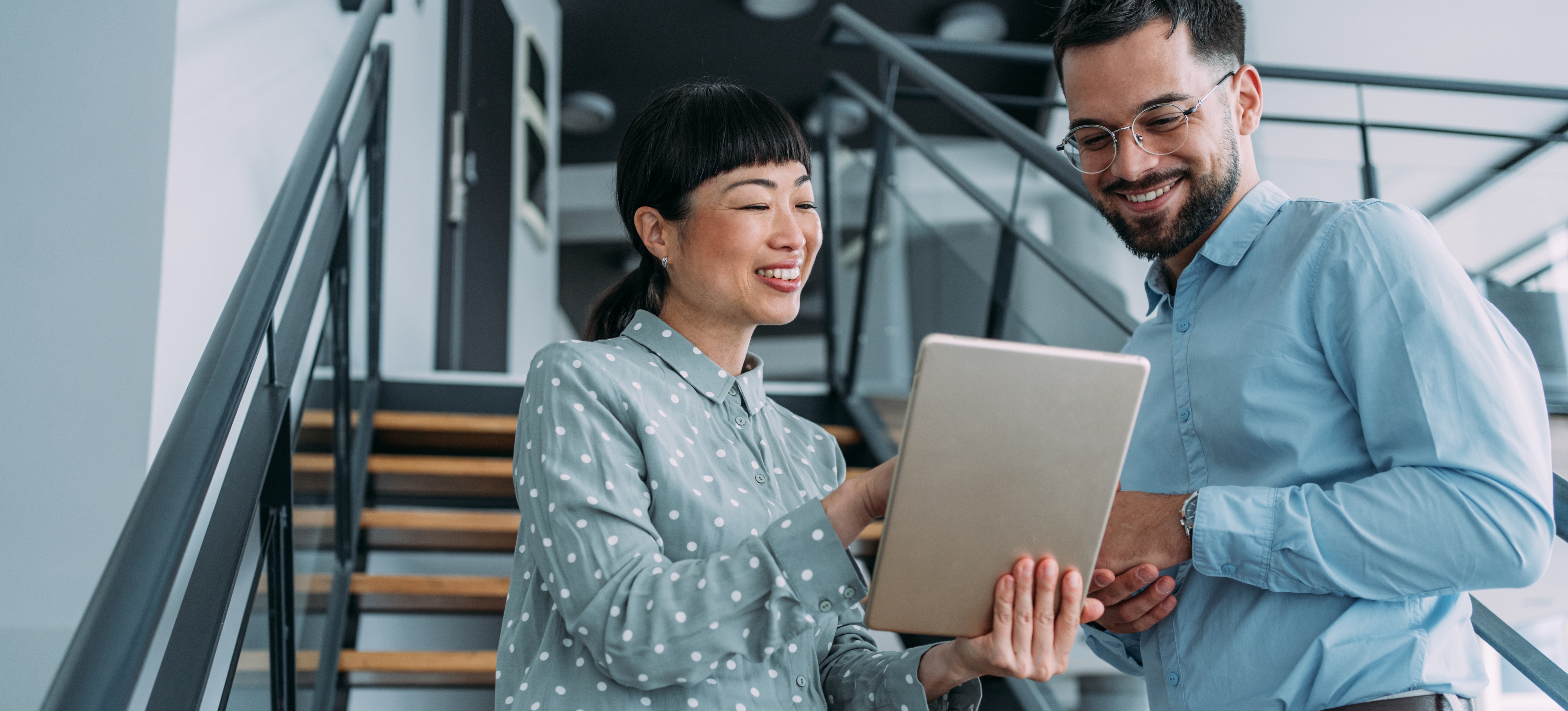 [Featured Image] Two social media strategists meet in front of a stairway in their office building and look over a campaign on a tablet. 
