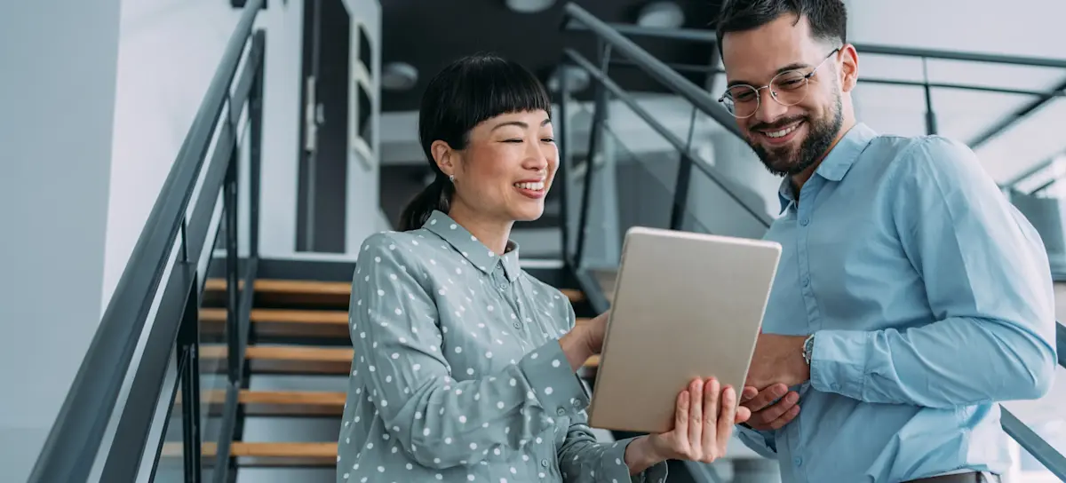 [Featured Image] Two social media strategists meet in front of a stairway in their office building and look over a campaign on a tablet. 
