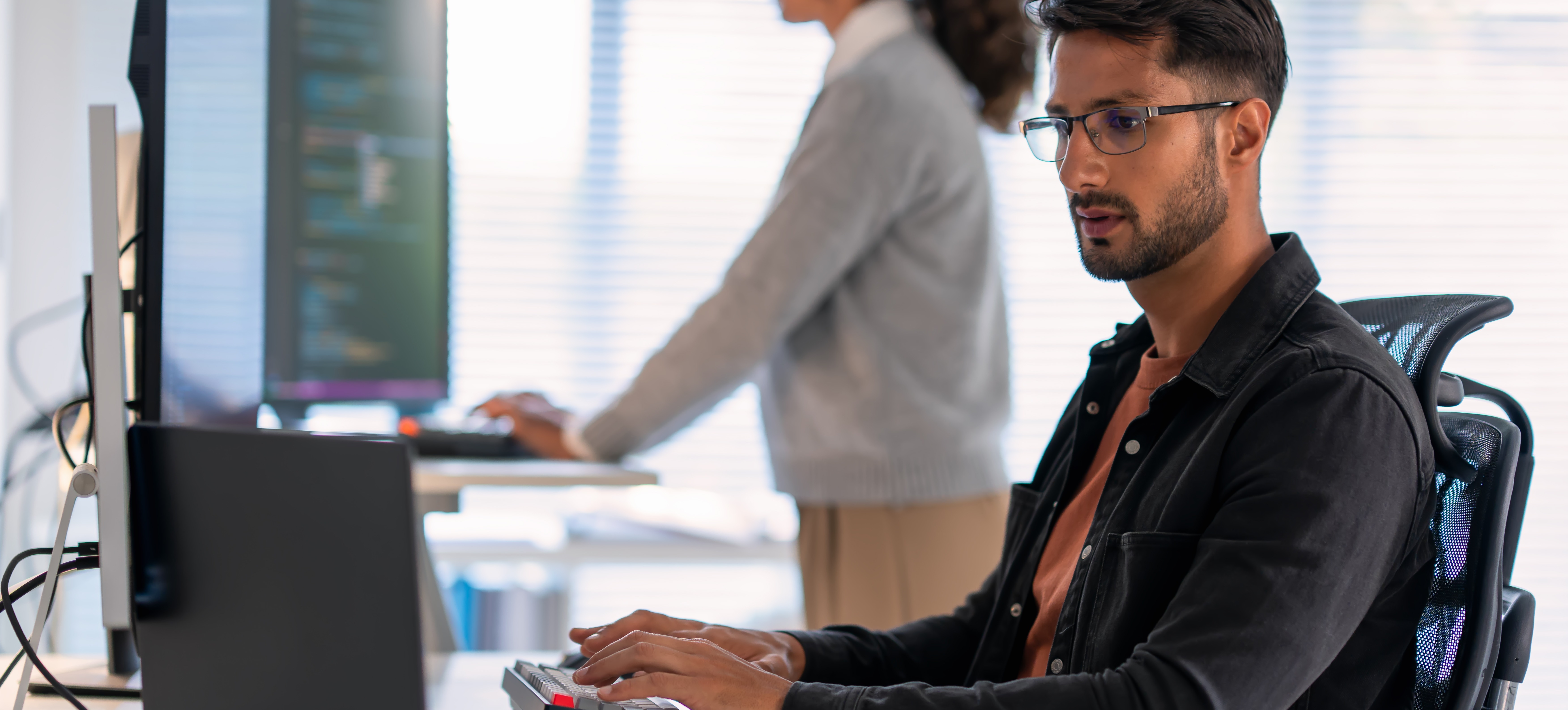 [Featured Image] A programmer sits at a computer and uses R programming skills while a colleague stands at a computer desk in the background.
