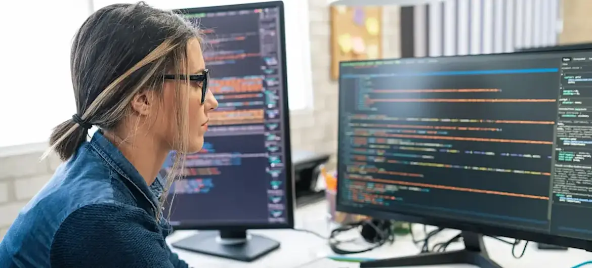 [Featured Image] A network automation engineer sitting at a desk programming and working on two computer screens.
