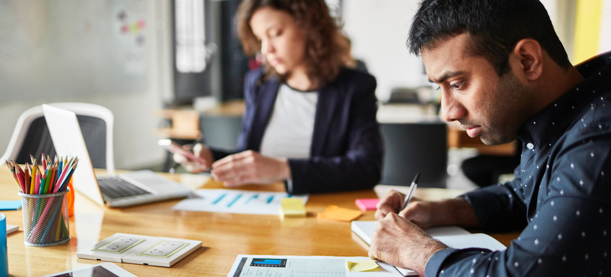 [Featured Image] Two people working at a desk, analyzing notes from a business report.