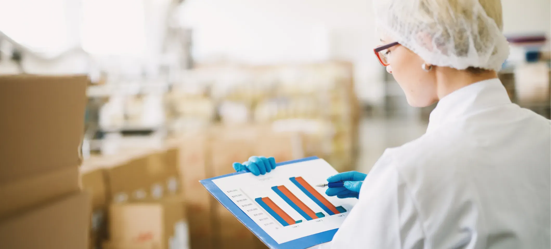 [Featured Image] A warehouse worker dressed in a white lab coat and wearing a white hair net and blue latex gloves studies a histogram on a clipboard.
