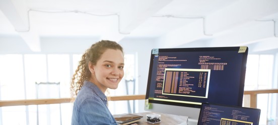 [Featured image] A female audio engineer, wearing a blue denim shirt is working with her laptop and desktop computers that are situated on a brown desk.