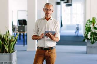 [Featured image] A manager in a white short and glasses stands in front of a white columned room holding a tablet.