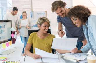 [Featured image]  Three data analysts review data on several report sheets in a large white room with two other analysts in the background