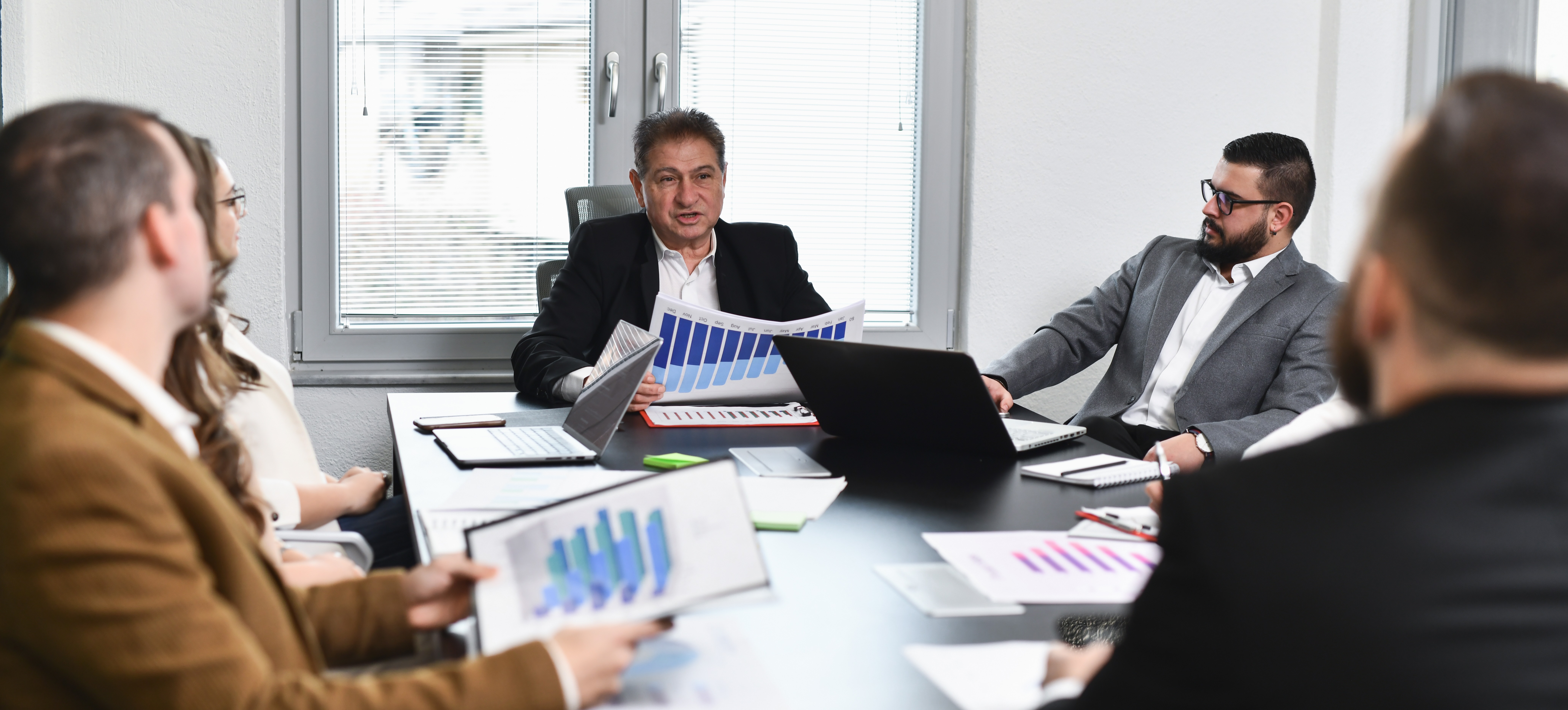 [Featured Image] A sales analyst holding bar charts meets in a boardroom with C-level executives to discuss quarterly sales.
