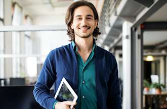 A data analyst in a blue sweater stands in front of his workstation holding a tablet