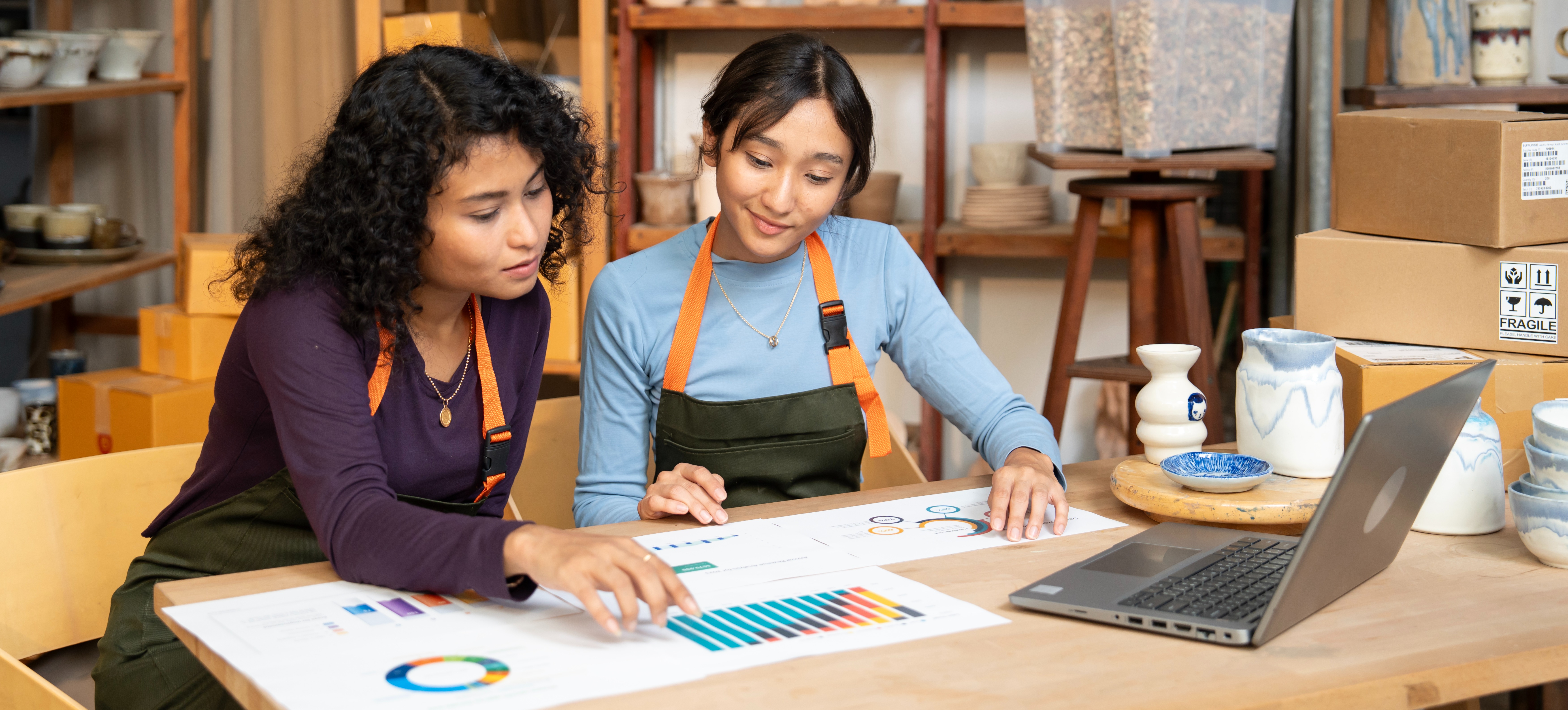 [Featured image]: Two small business owners are sitting at a table, surrounded by products as they analyze data charts provided by a data analyst.

