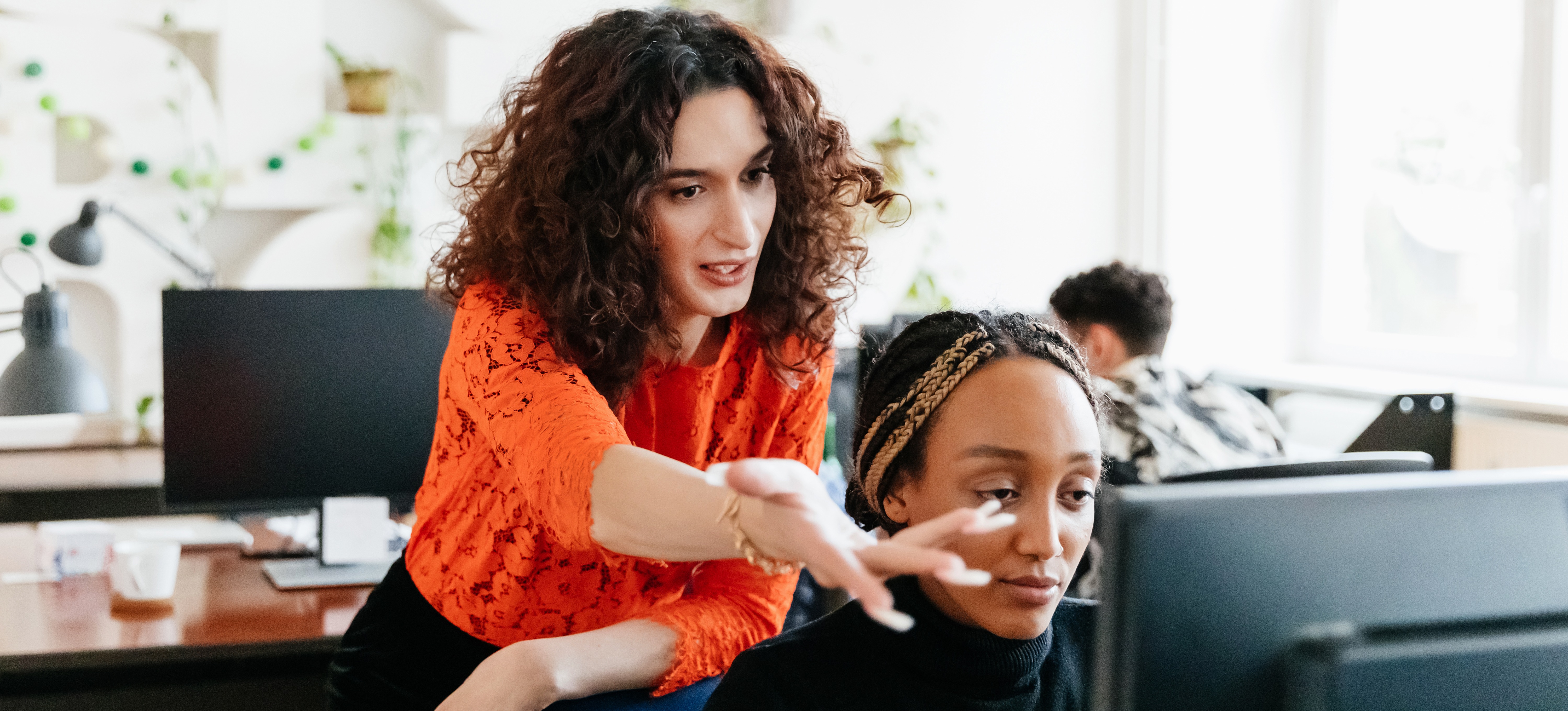 [Featured Image] Two colleagues review their programmatic ad buying strategy on one of their desktop computers in a sunny office. 
