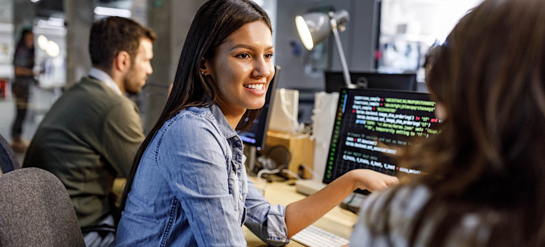 [Featured Image] Two software developers discuss unit testing in front of a computer screen in an office setting with other colleagues in the background.
