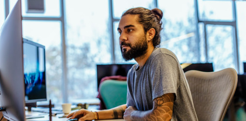 [Featured image] A computer technician works at a dual-screen workstation in an office with windows behind him. 