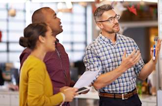 [Featured Image] A technical project manager walks a team through a project plan on a whiteboard.