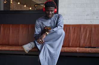 A man wearing red headphones sits on a leather bench listening to a podcast on his phone.