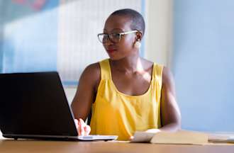 [Featured image] A data analyst wearing a yellow tank top and eyeglasses sits in front of a laptop in an office writing a statistical analysis program.