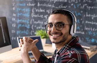 [Featured Image] A programmer wearing a plaid shirt, glasses, and headphones sits at a desk with a laptop and an external monitor with code running down the screen.