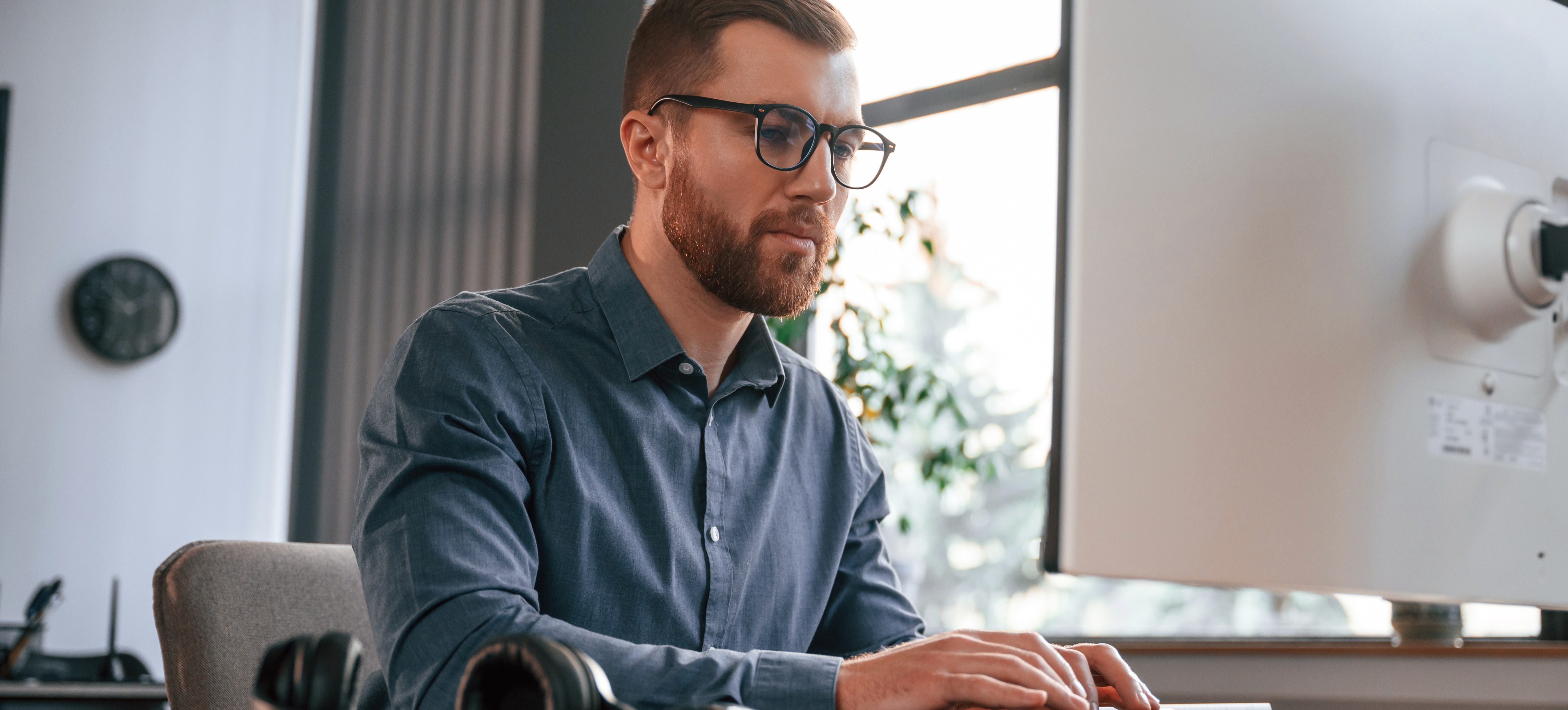 [Featured Image] An AI prompt engineer works with generative AI on his computer in an office near a sunny window.
