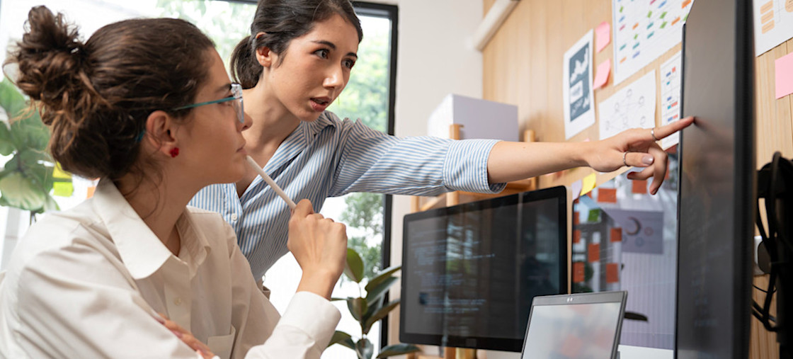 [Featured Image] A cybersecurity professional is sitting at a desk with two monitors and a laptop while a colleague stands next to them, pointing at one of the monitors. 
