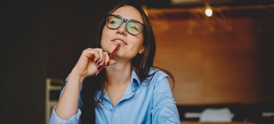 [Featured Image] A person is sitting at a desk pondering into the distance while holding a pen to their mouth.