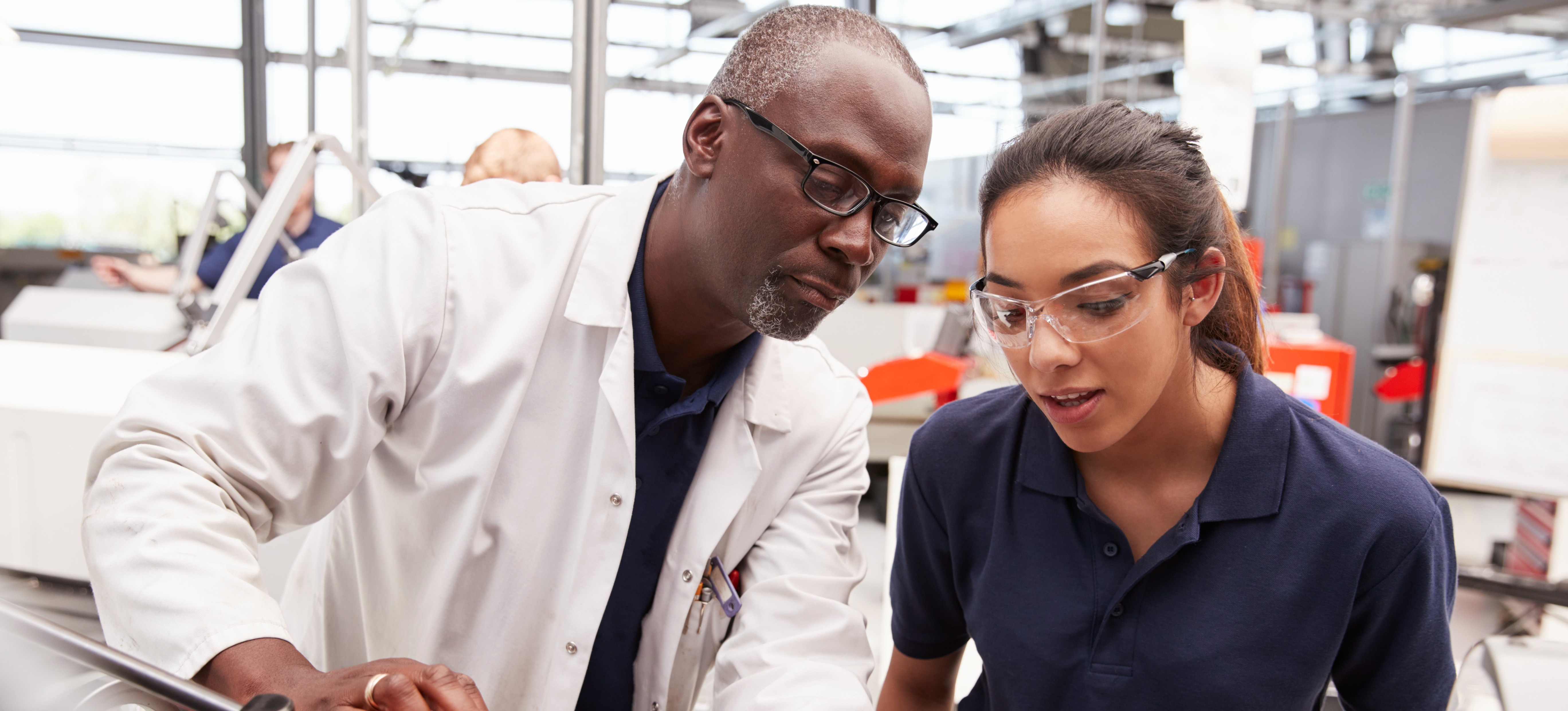 [Featured Image] After learning what is an externship, a young person in safety glasses shadows an engineer in a white coat, as they show them equipment in their workspace.

