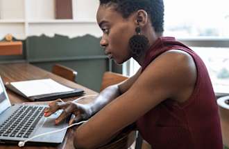 [Featured Image] A person in a maroon shirt sits at a wooden table and works on their LinkedIn profile on a laptop. 