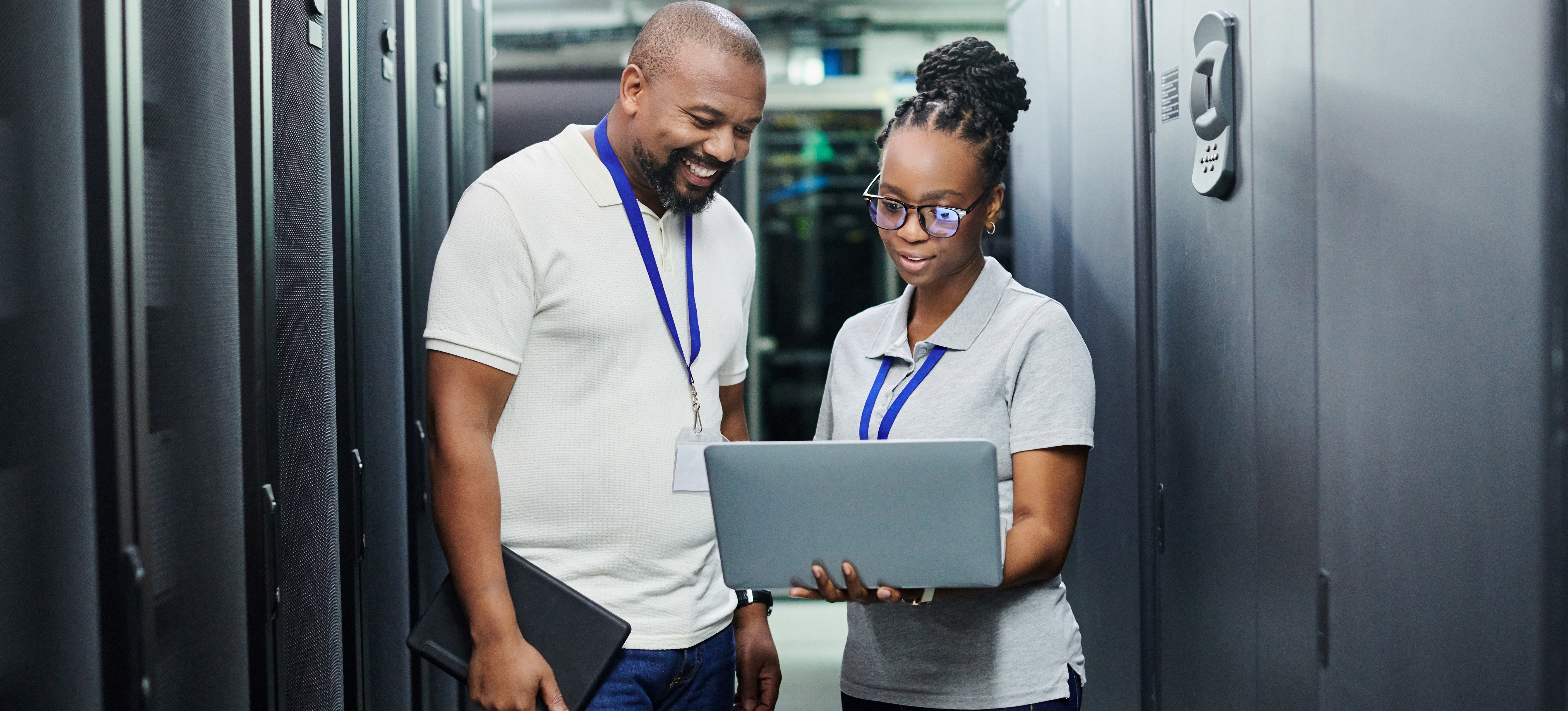 [Featured Image] Network administrators collaborating in an office environment, with one holding a computer while discussing the network administrator certification they consider pursuing. 

