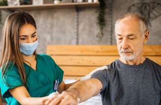 [Featured Image]:  A female home care aide, wearing a green uniform and face covering, is taking care of a male patient at his home.