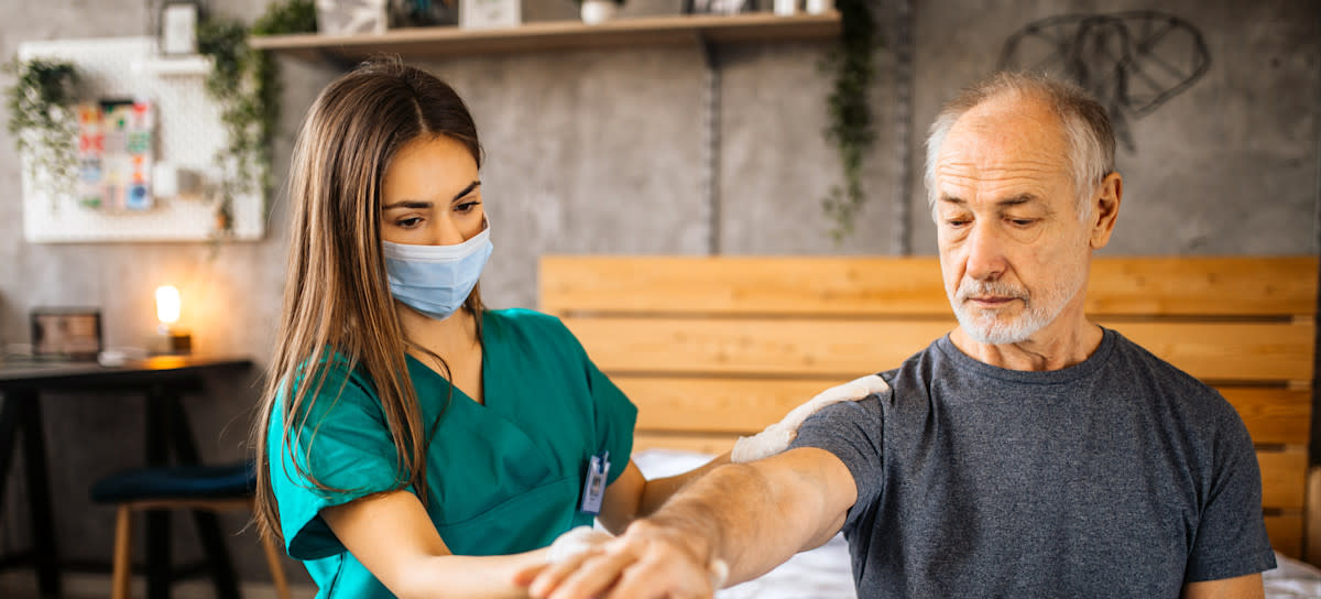 [Featured Image]:  A home care aide, wearing a green uniform and face covering, is taking care of a patient at their home.